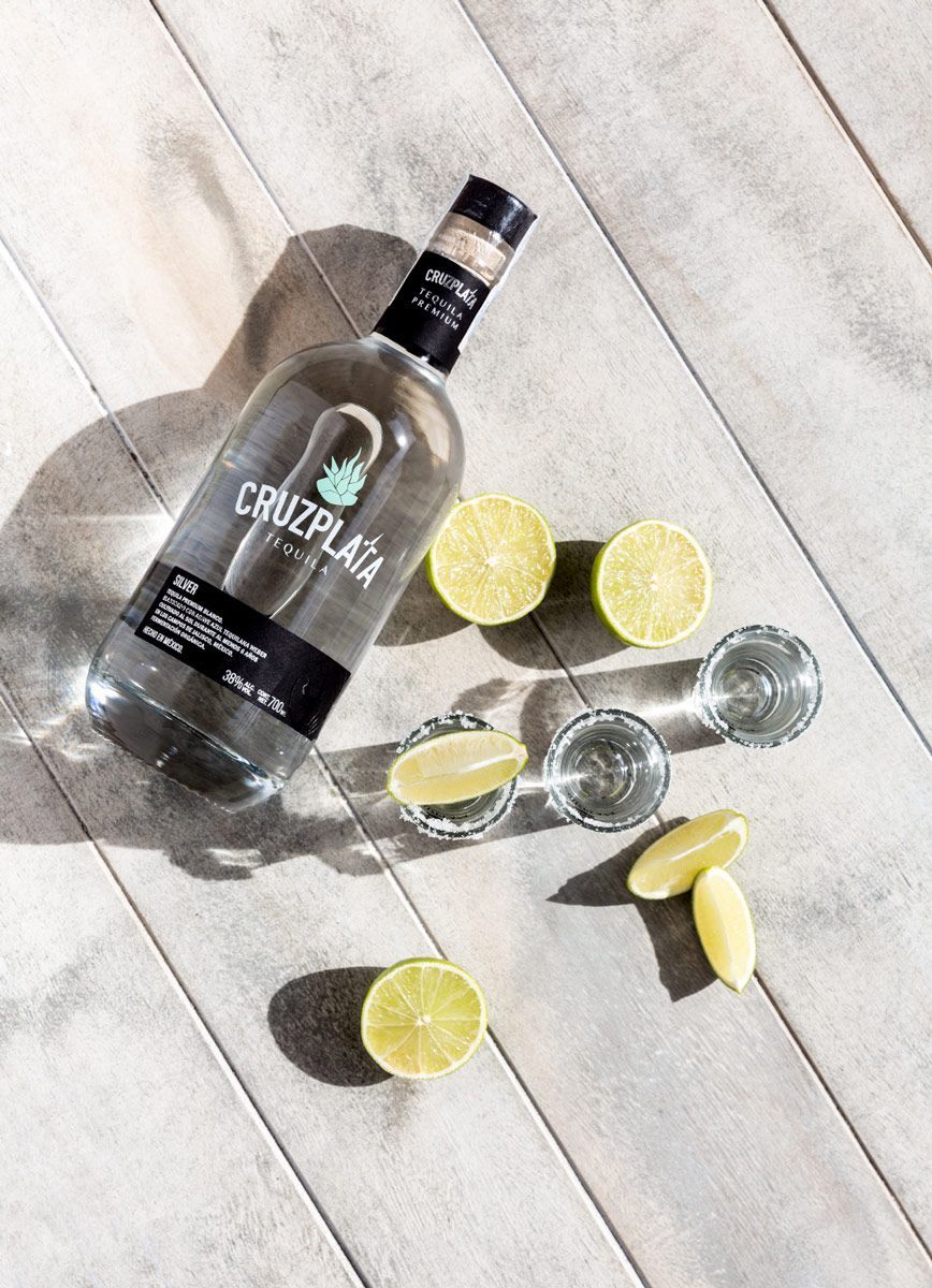 Una botella de vodka Cruz Plata se encuentra sobre una mesa de madera junto a rodajas de lima y vasos de chupito.