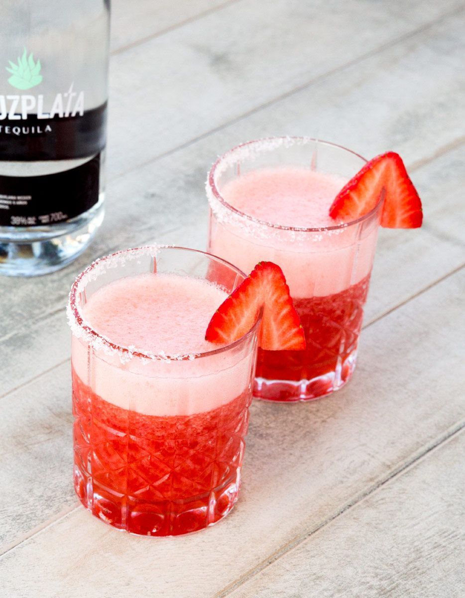 Dos vasos de margarita de fresa junto a una botella de tequila