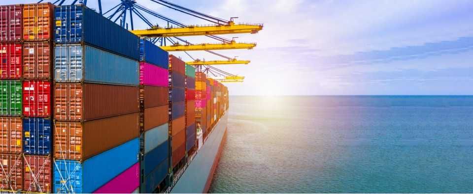 Kleurrijke zeecontainers opgestapeld op een vrachtschip op zee, naast havenkranen onder een heldere hemel.