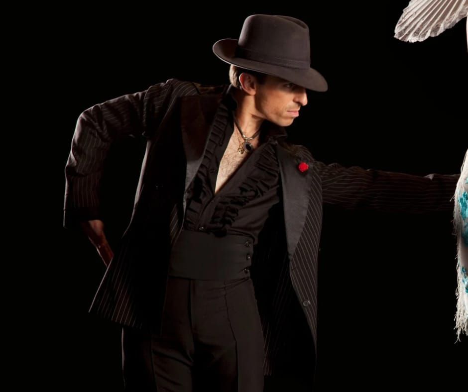 Hombre con traje negro y sombrero fedora bailando con una pluma de pavo real, sobre un fondo negro.