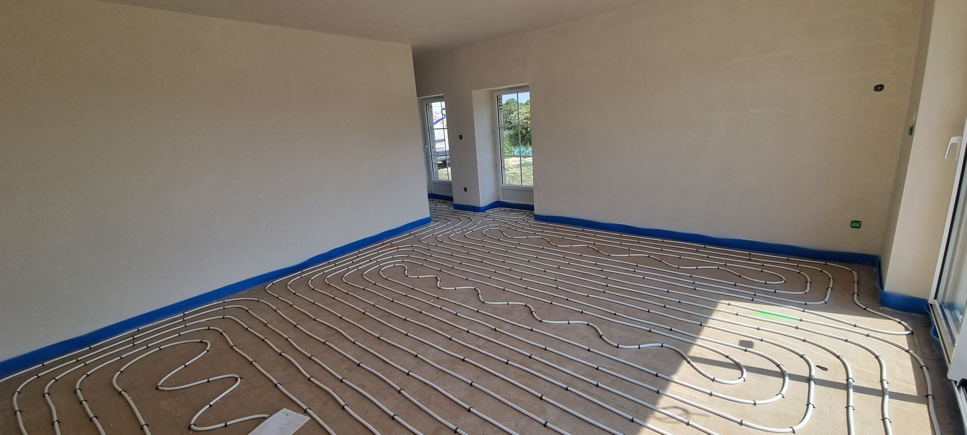 Pièce équipée d'un chauffage au sol, délimitée par un ruban bleu. Murs peints en blanc cassé. La lumière naturelle y pénètre abondamment.