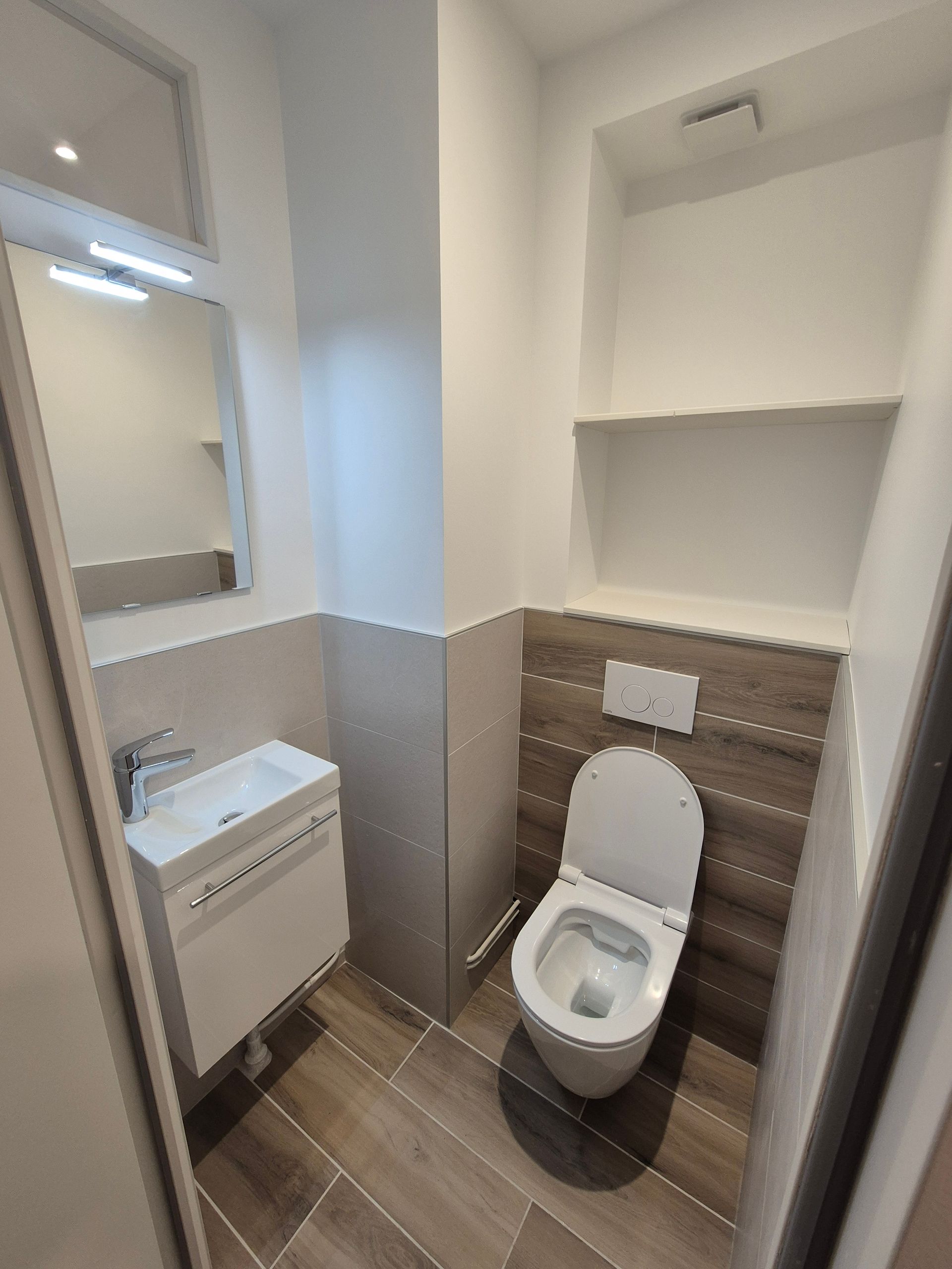 Petite salle de bains moderne avec toilettes, lavabo, miroir et étagères. Couleurs neutres, touches de bois.