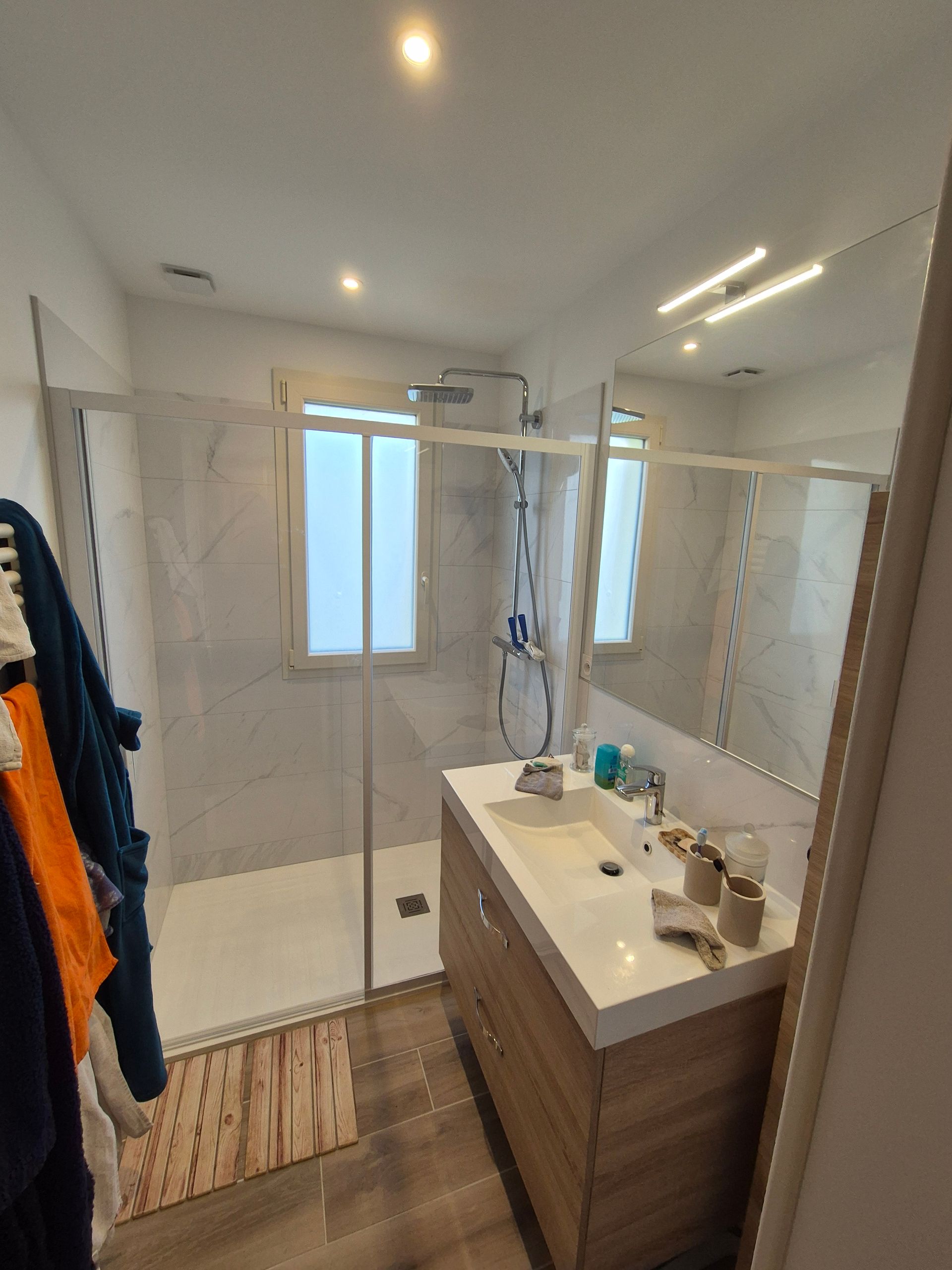 Salle de bains moderne avec douche en verre, meuble-lavabo en bois et murs carrelés imitation marbre.