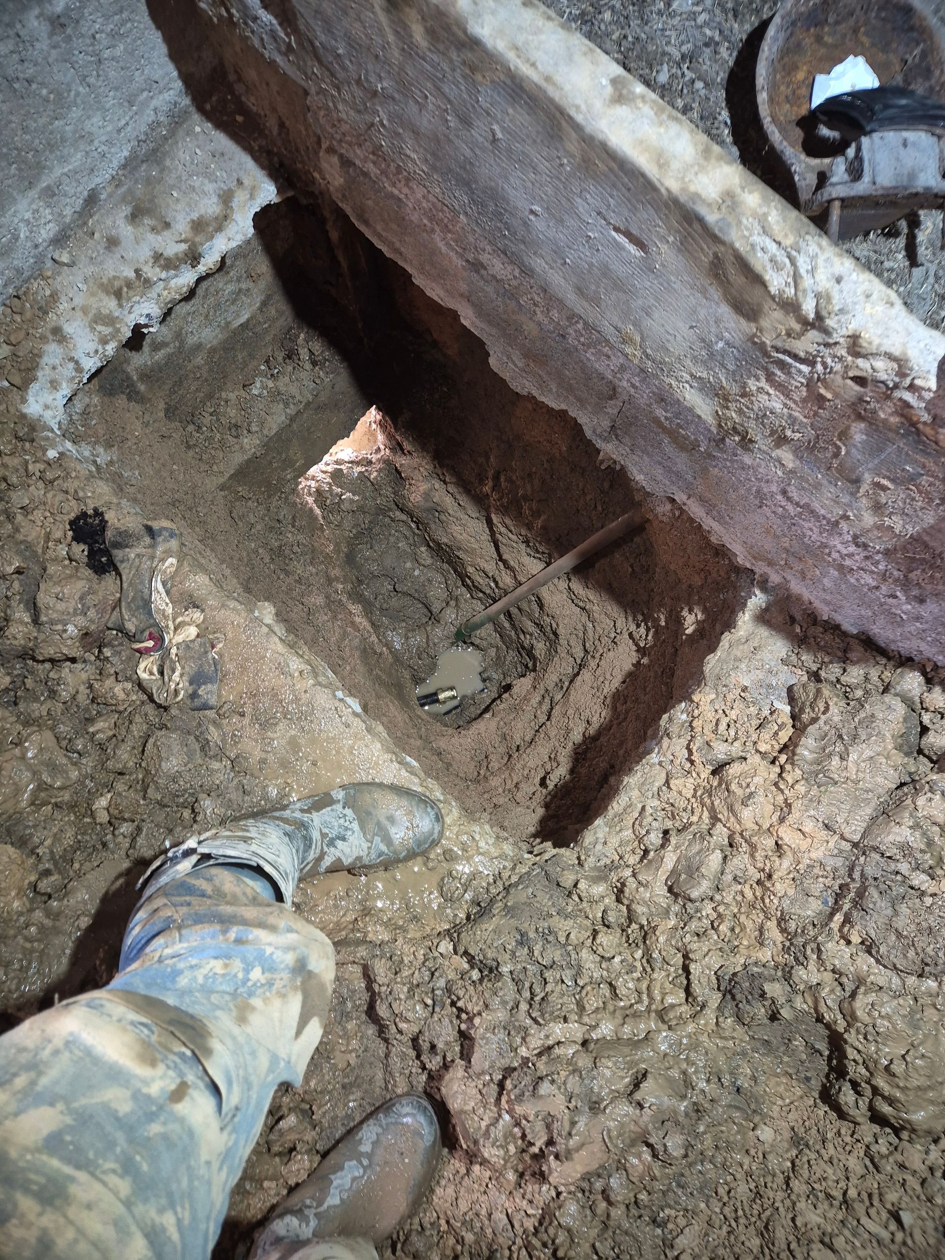 Les bottes et les jambes boueuses d'une personne à l'intérieur d'un trou creusé dans le sol sous une poutre en bois.