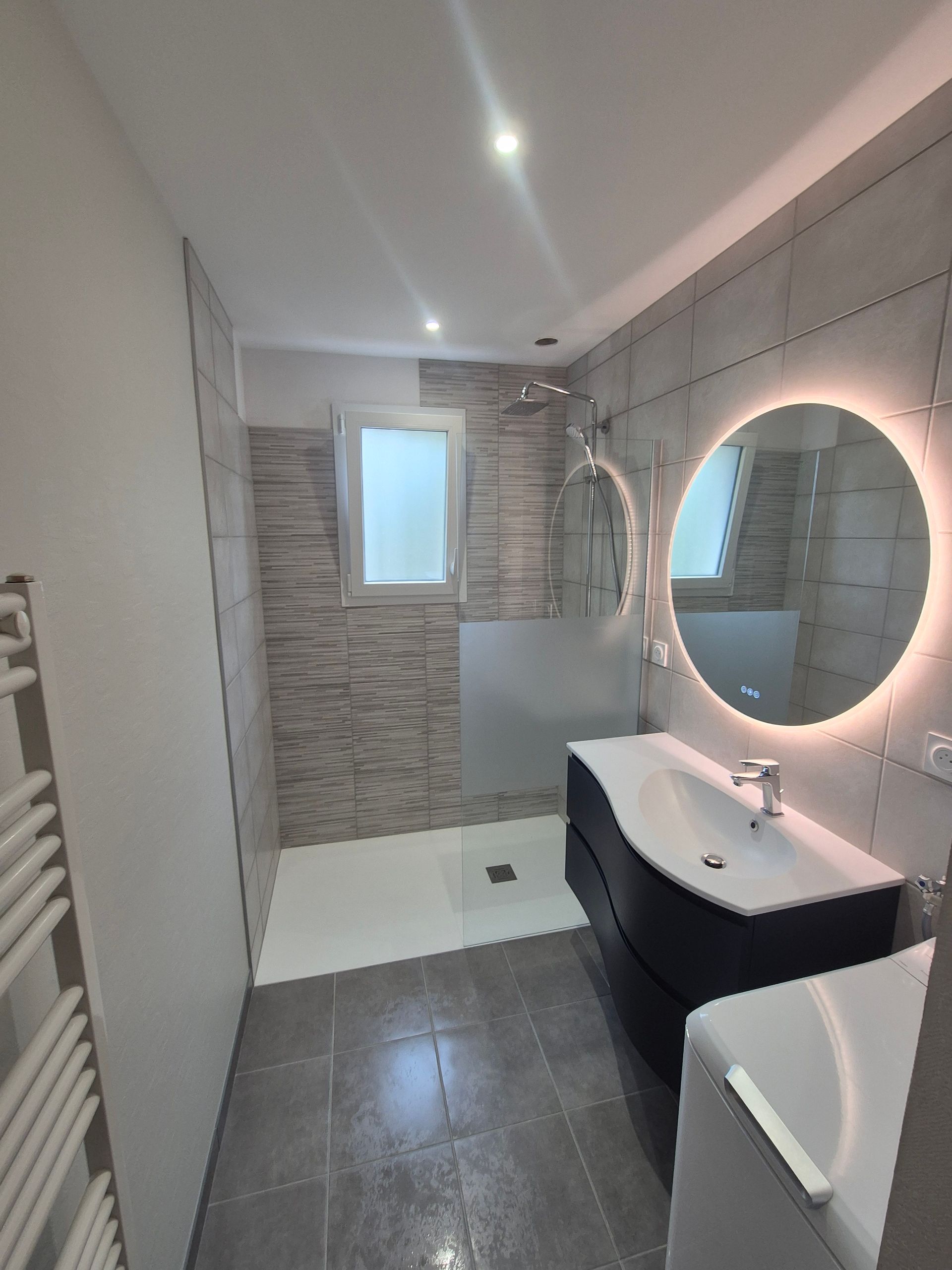 Salle de bains moderne avec carrelage gris, douche, lavabo et miroir rond.