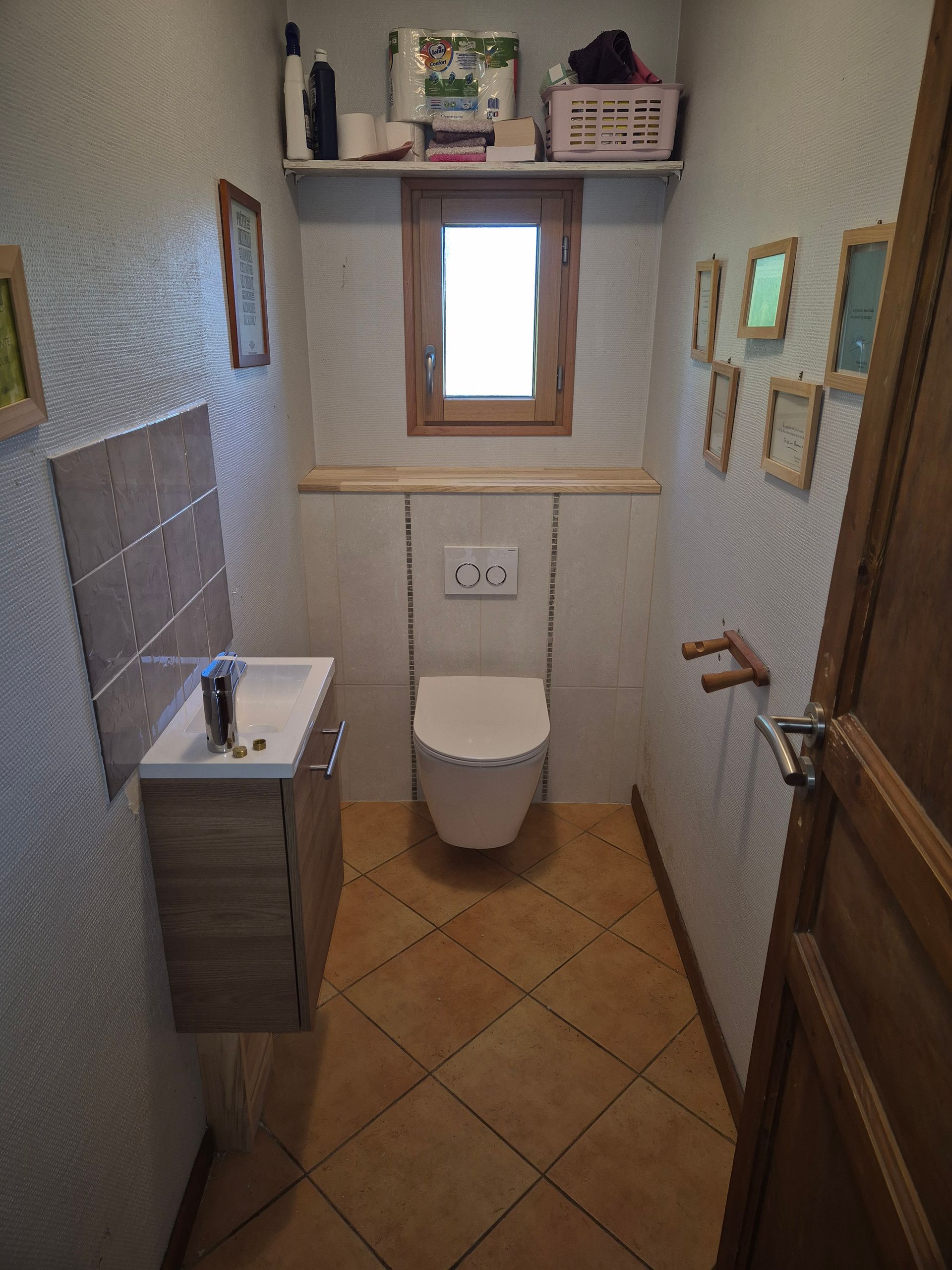 Petite salle de bains avec toilettes, lavabo et porte en bois ; sol carrelé beige et murs de couleur claire.
