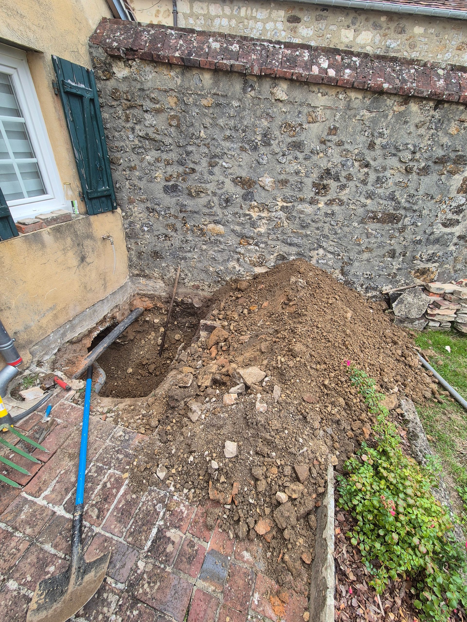 Trou creusé près d'un bâtiment en pierre, avec un tas de terre à côté. Pelle à manche bleu sur un sol en briques.