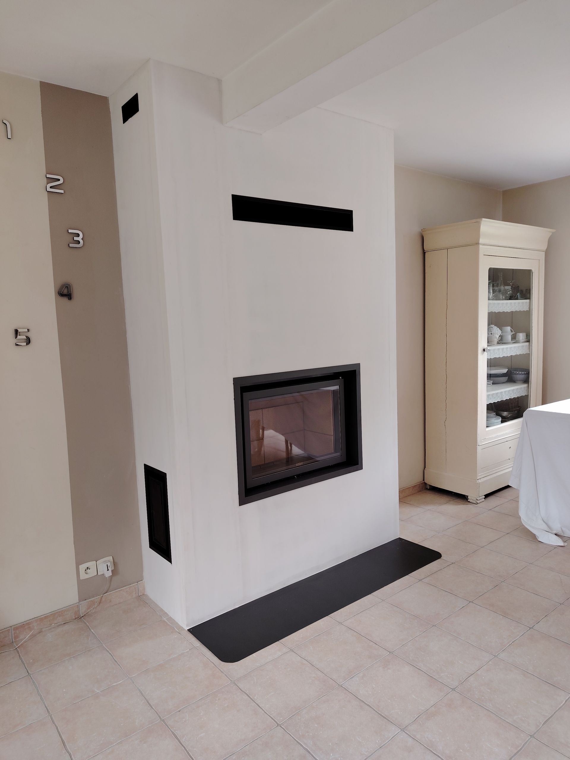 Cheminée blanche à encadrement noir, entourée de murs blanc cassé. Grilles d'aération noires et foyer sombre.