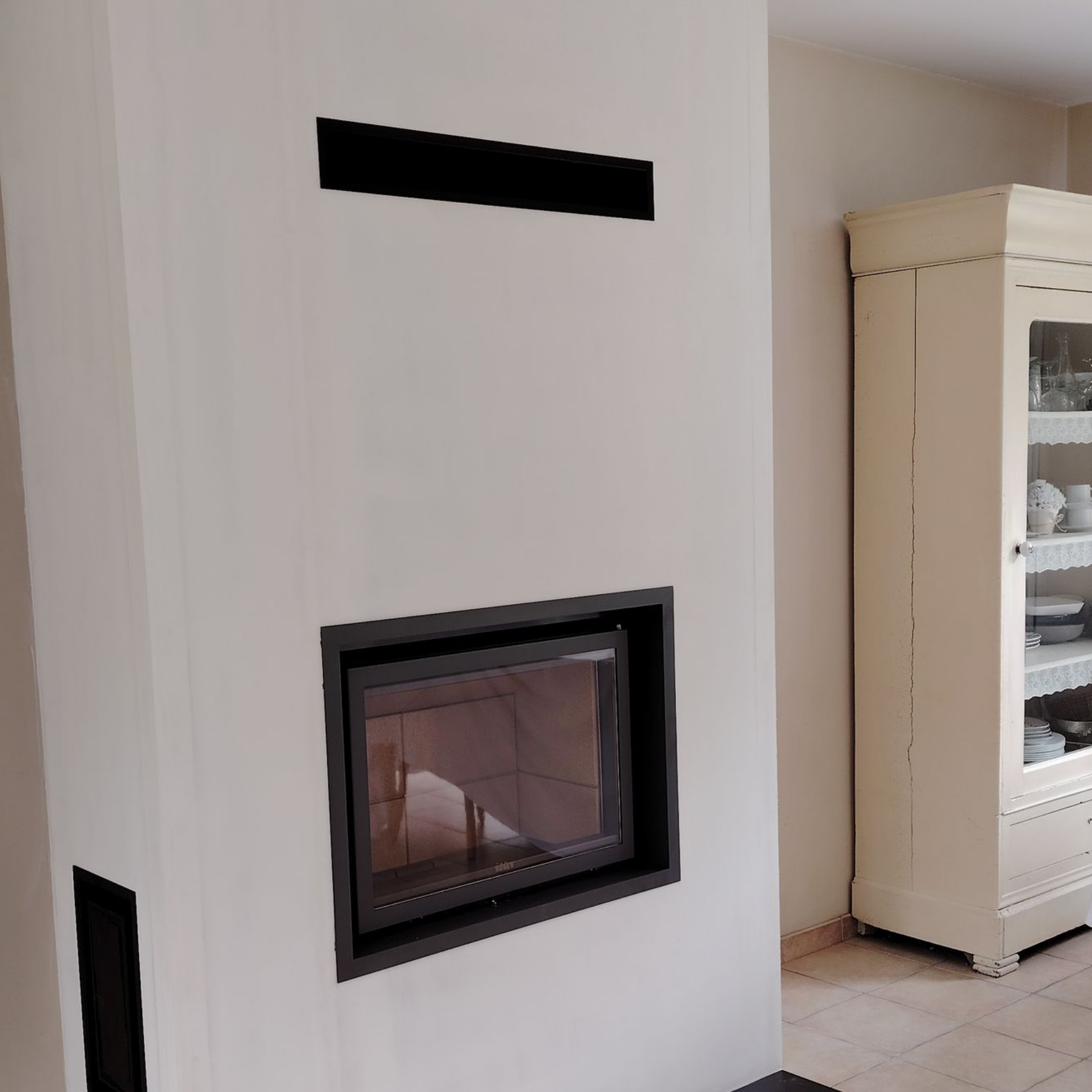 Cheminée avec cadre et conduit noirs sur un mur blanc ; meuble blanc cassé à droite.