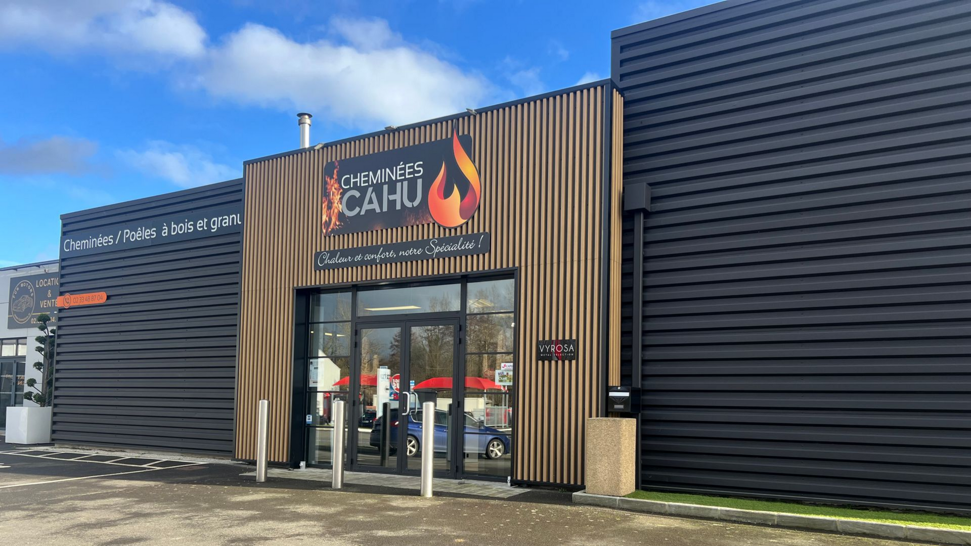 Extérieur du magasin Cheminées CAHU, avec un logo au-dessus de la porte vitrée. Bardage noir et panneaux de bois.