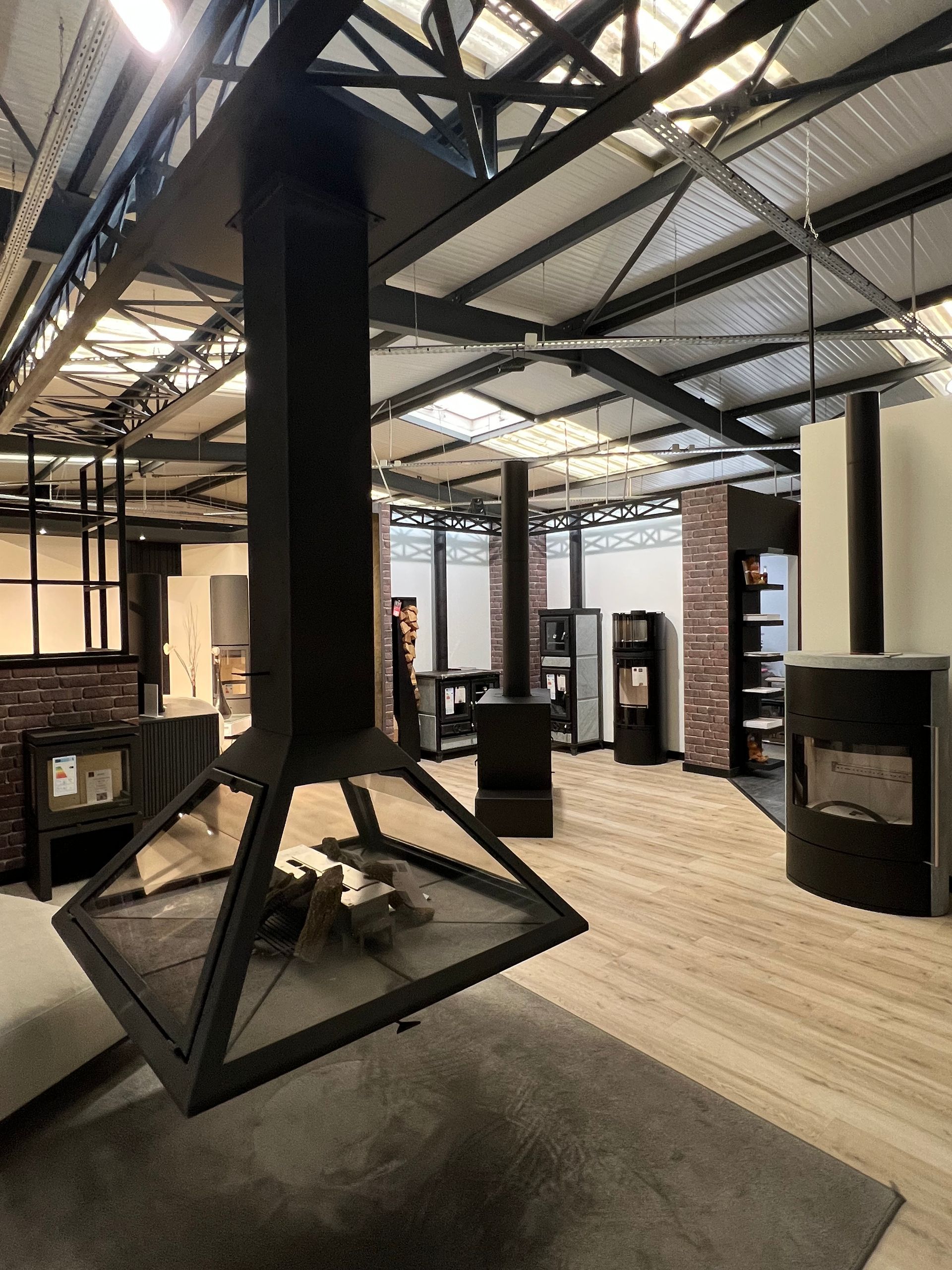 Cheminée noire et moderne suspendue dans une salle d'exposition, parquet, murs en briques, poutres métalliques.