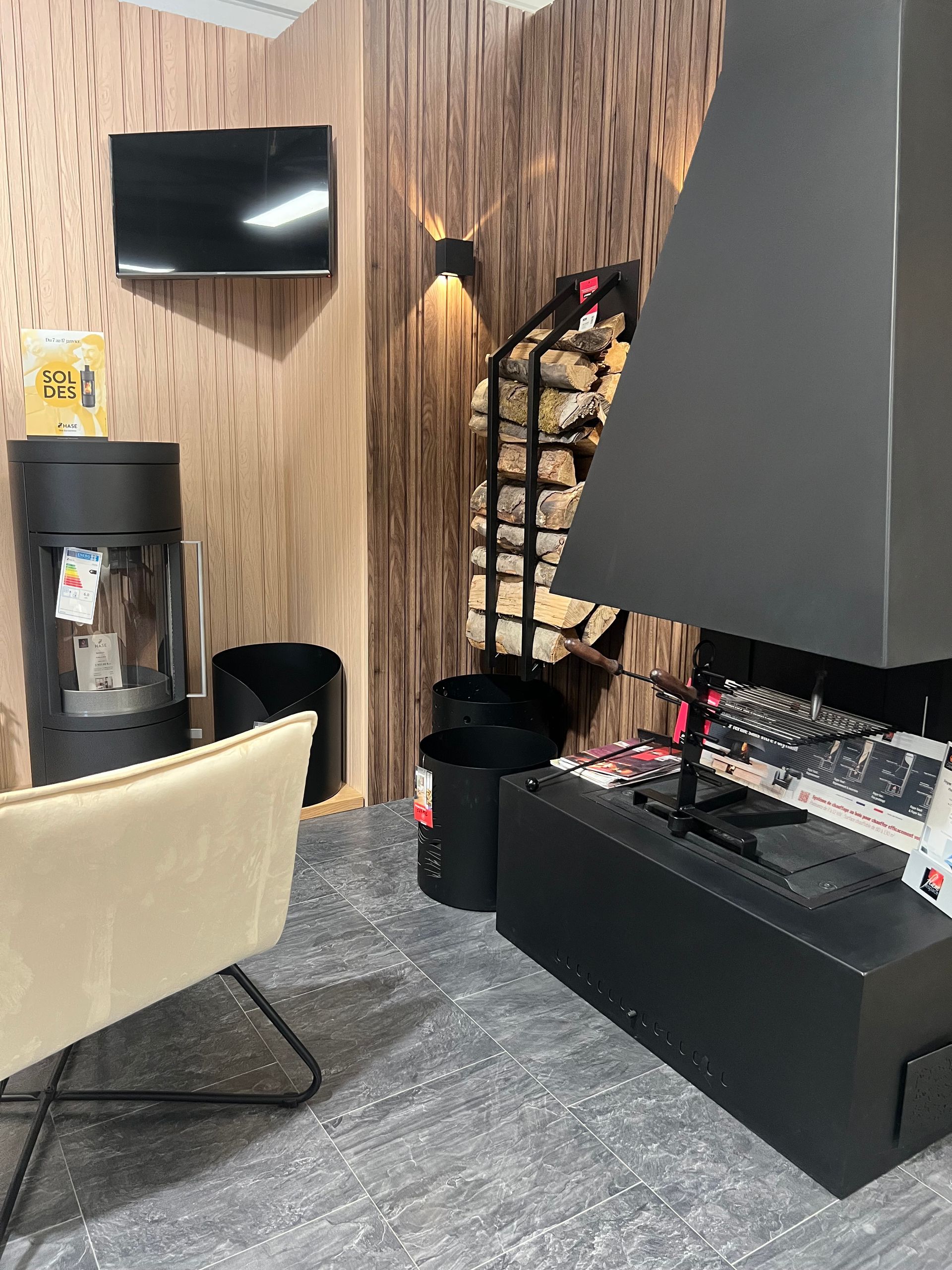 Aménagement d'un salon moderne comprenant une cheminée, un porte-bûches, une télévision et un fauteuil ; tons neutres.