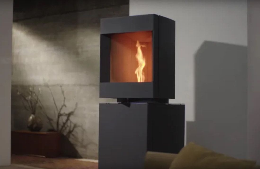 Cheminée moderne noire à flammes orange. Elle repose sur un socle sombre adossé à un mur en béton.