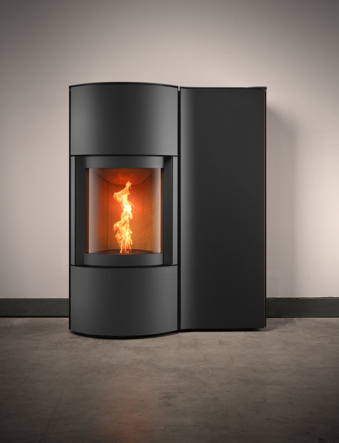 Poêle à granulés cylindrique noir avec vitre laissant apparaître les flammes sur un mur clair et un sol gris.