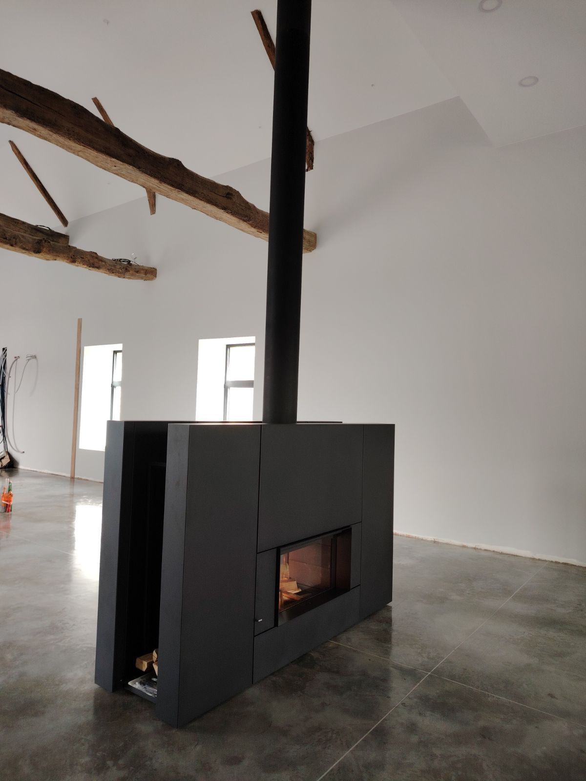 Une cheminée moderne noire suspendue au plafond dans une pièce aux murs blancs et aux poutres apparentes.