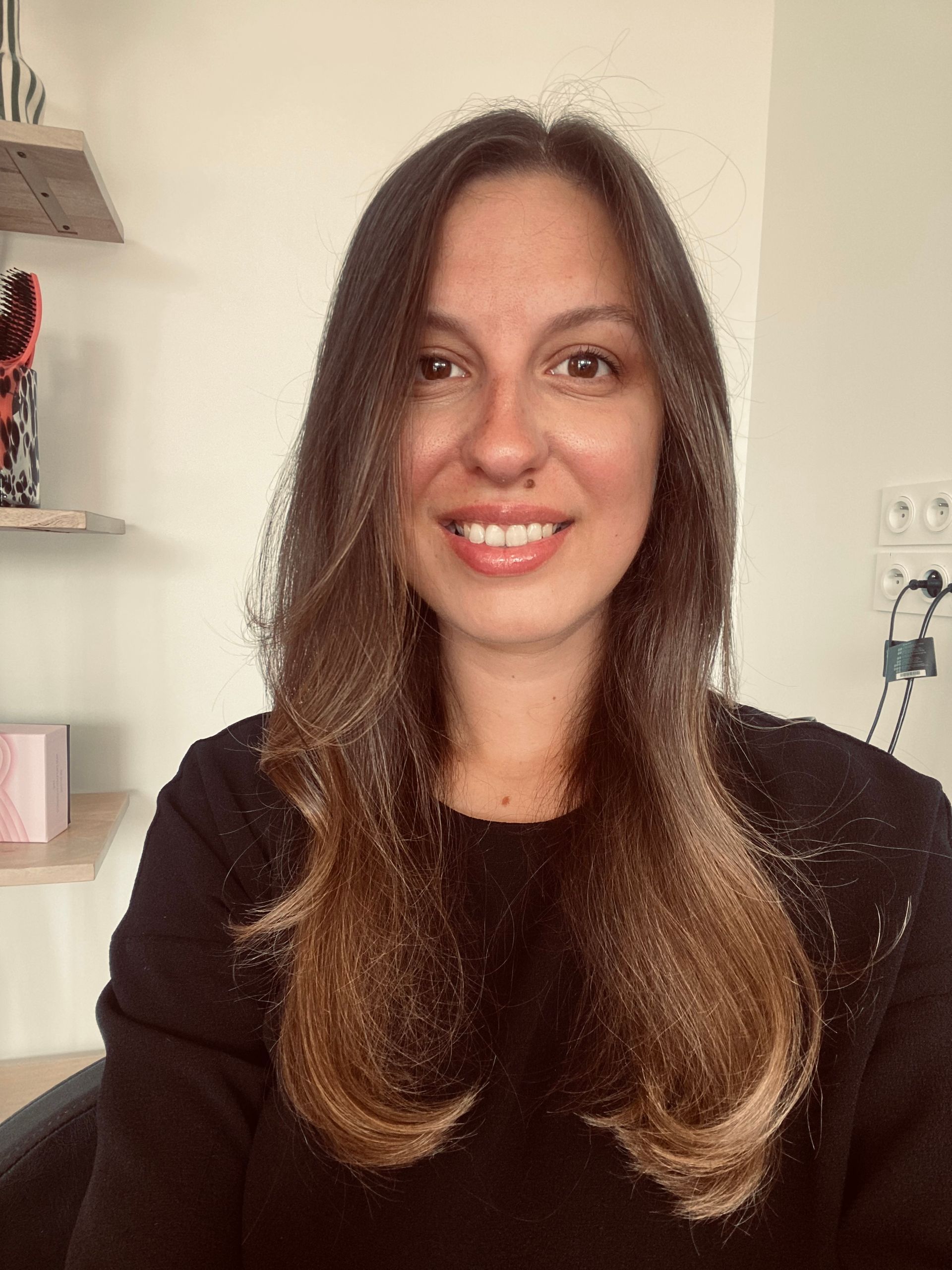 Femme aux longs cheveux bruns souriant à l'objectif devant un mur blanc.