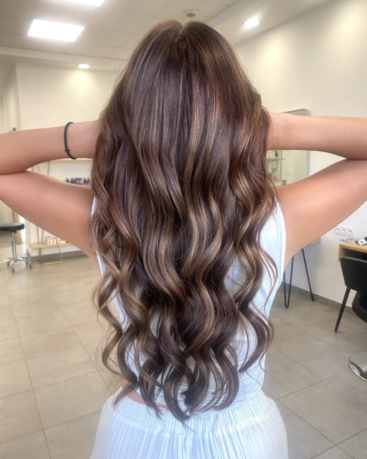 Cheveux longs, bruns et ondulés avec des mèches, vus de dos, dans un salon de coiffure bien éclairé.