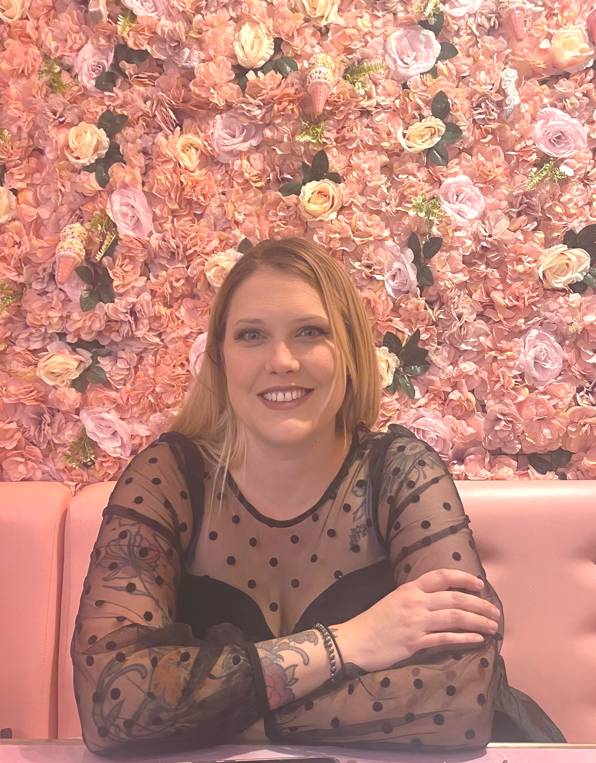 Une femme assise, les bras croisés, devant un mur fleuri rose.