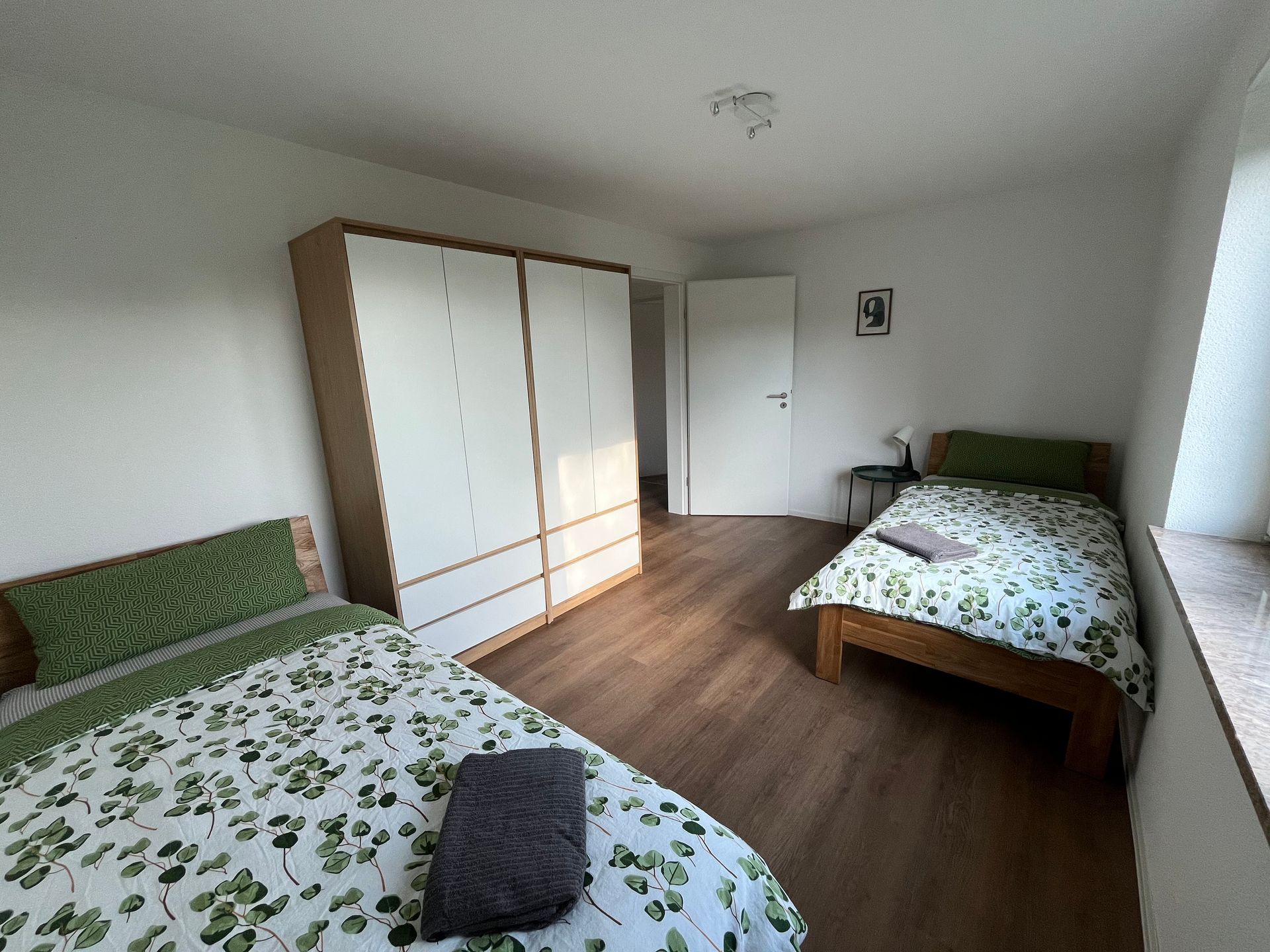 Ein Schlafzimmer mit zwei Betten, einem gemusterten Teppich, einer olivgrünen Wand und einem Fenster mit weißen Vorhängen.