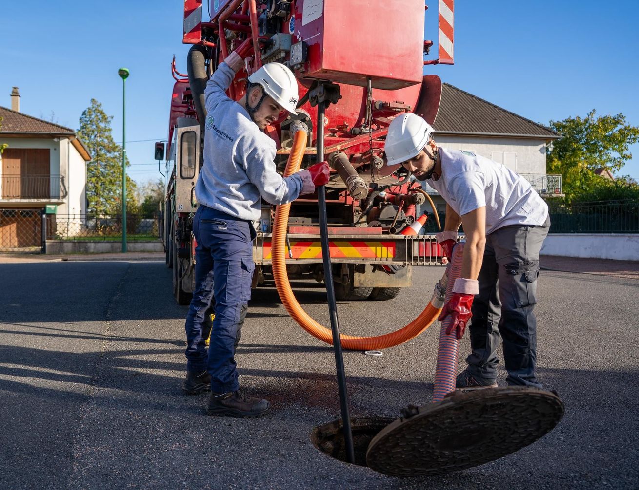 Deux ouvriers casqués réparent un nid-de-poule dans une rue, à côté d’un camion de travaux publics.