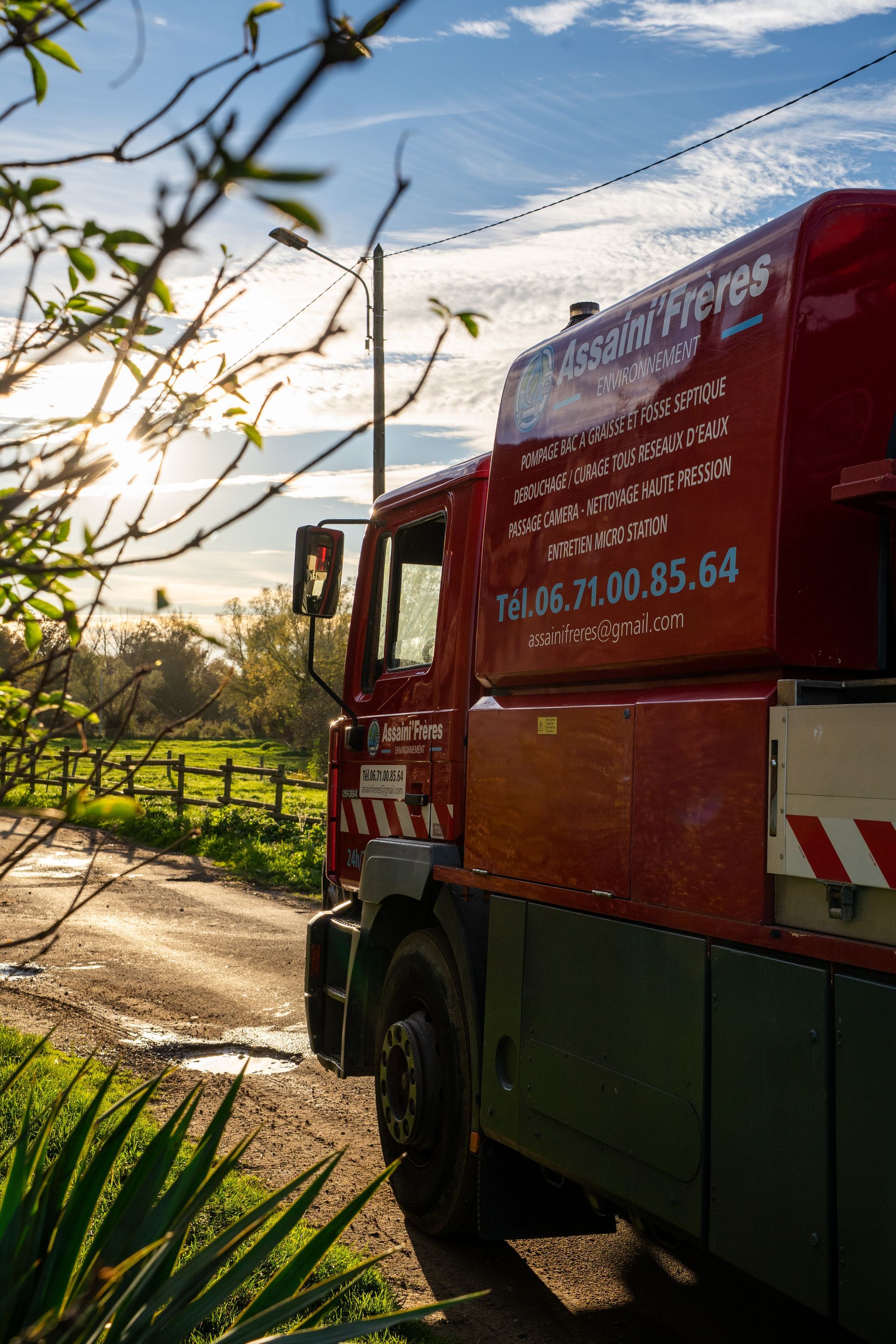 Camion rouge sur une route de campagne, près d’un champ ensoleillé et d’une clôture, sous un ciel bleu.