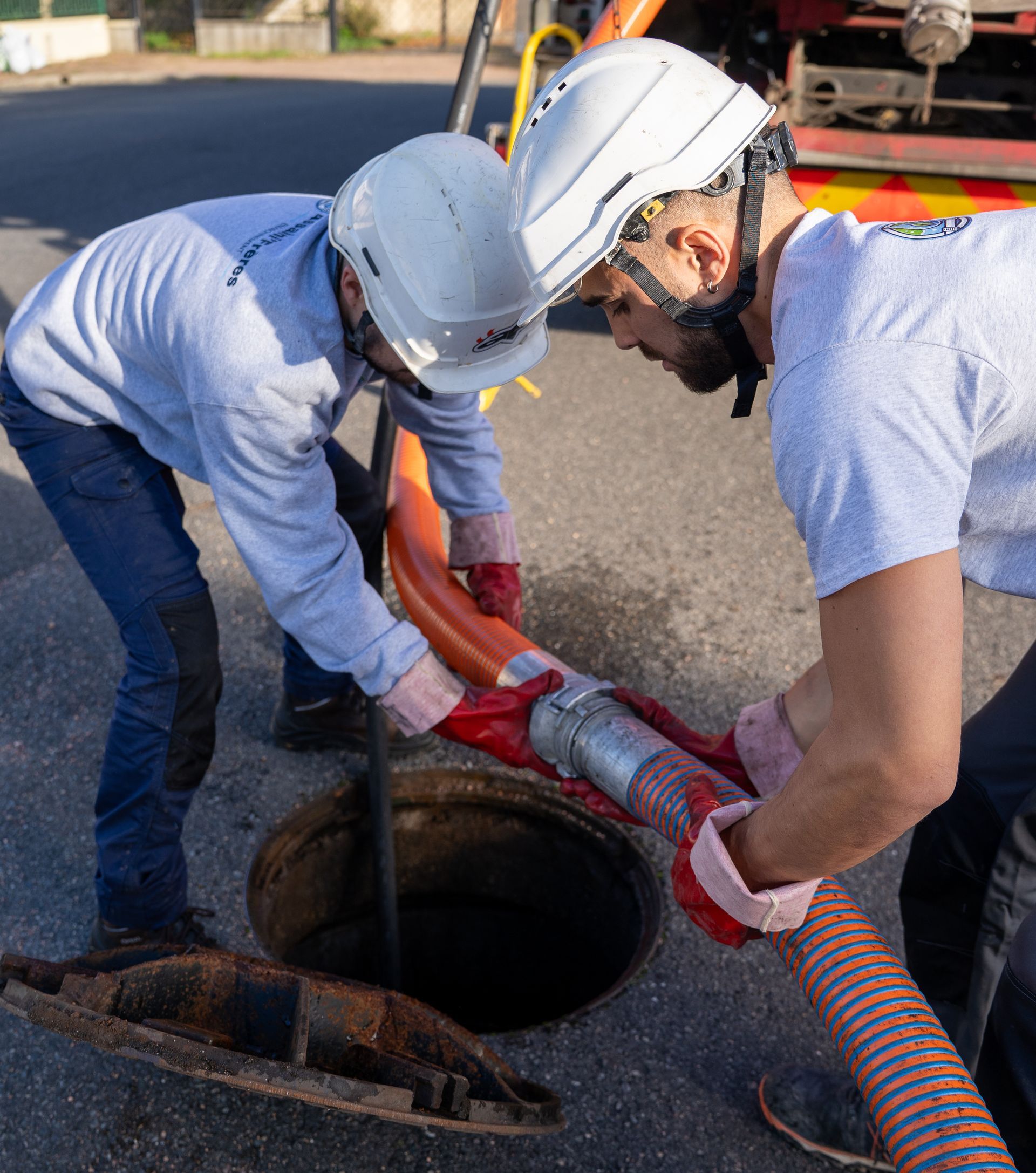 Deux ouvriers portant des casques de chantier tirent un tuyau rouge d’une bouche d’égout ouverte dans la rue.