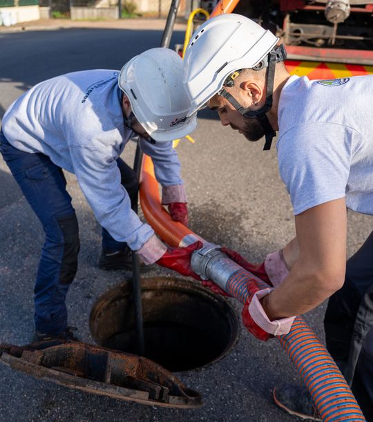 Deux ouvriers portant un casque utilisent un tuyau pour inspecter une bouche d’égout dans la rue.