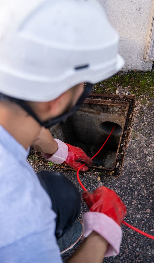 Un ouvrier portant un casque blanc manipule un câble rouge dans une trappe de service extérieure ouverte.