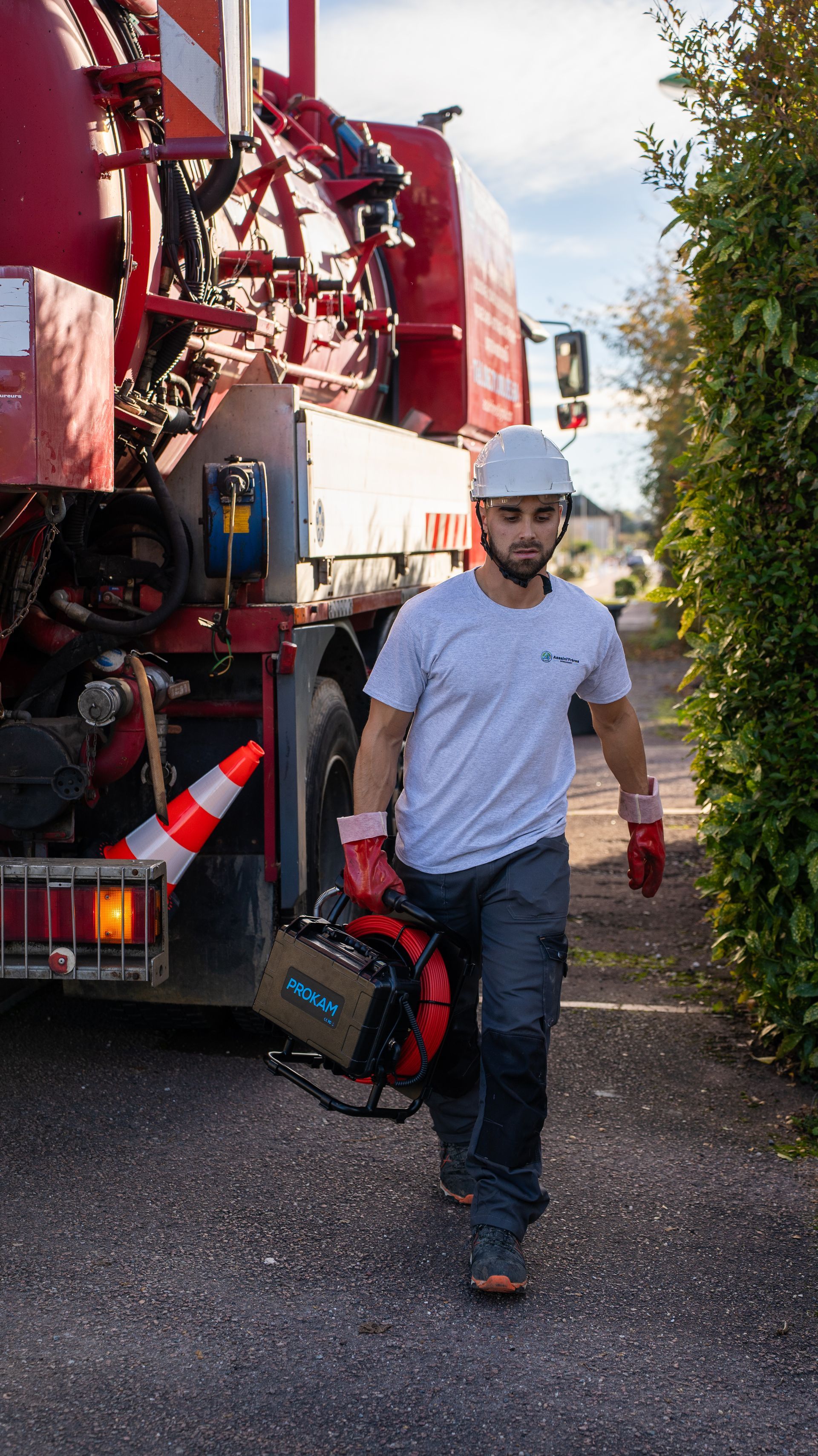 Un ouvrier portant un casque blanc marche à côté d’un camion rouge, le long d’une route goudronnée.
