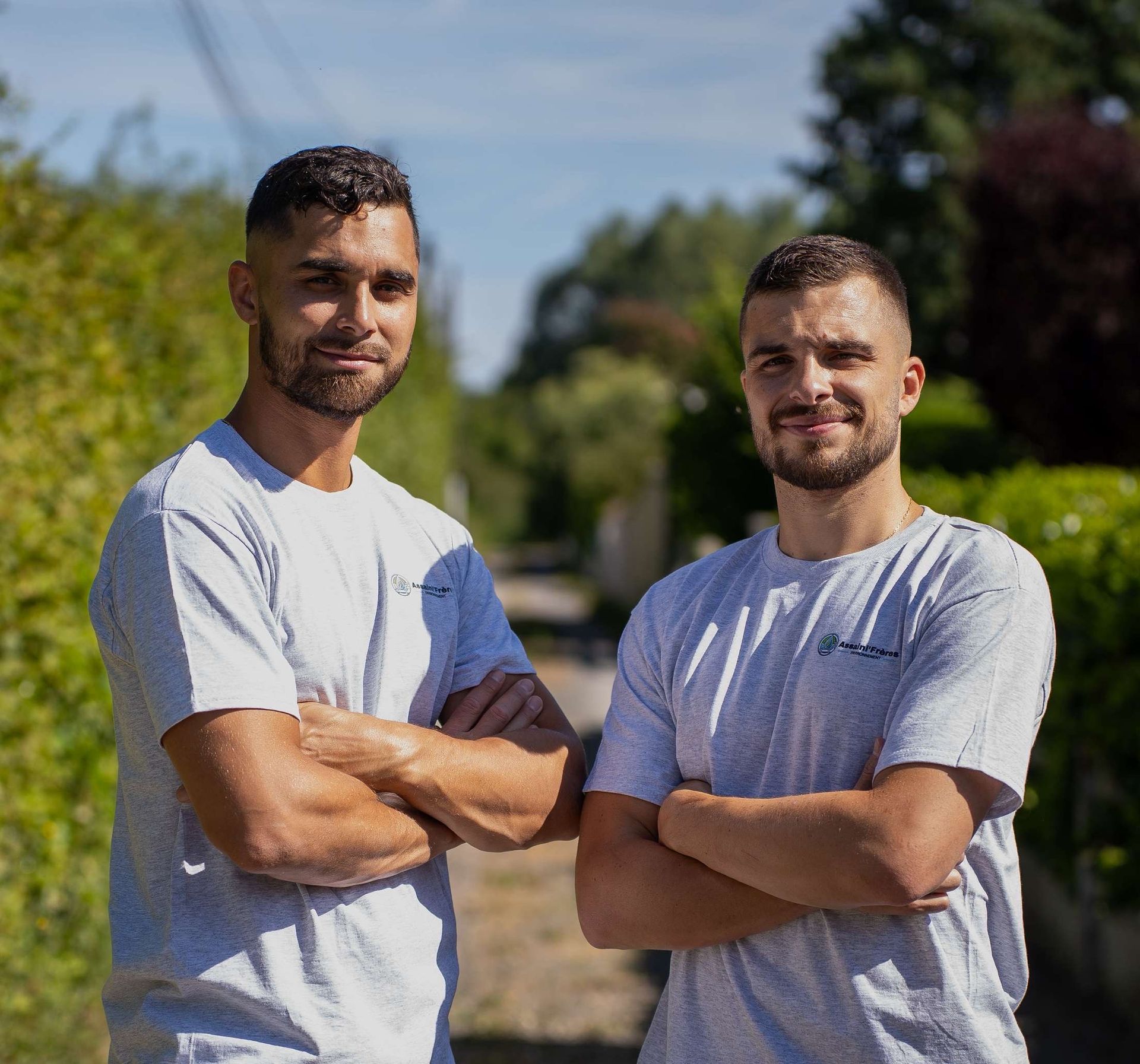 Deux hommes en T-shirts gris et blancs, debout dehors, les bras croisés et souriant au soleil.