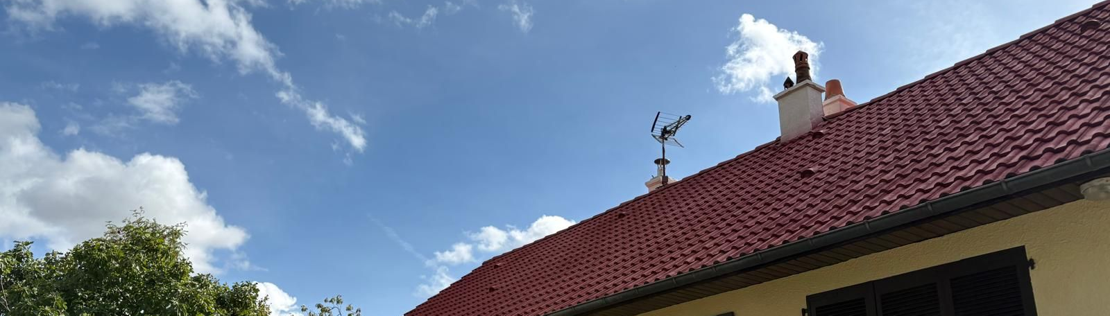 Le toit d'une maison aux tuiles rouges se détache sur un ciel bleu parsemé de nuages ​​blancs duveteux. Une antenne en forme de cigogne orne le toit.