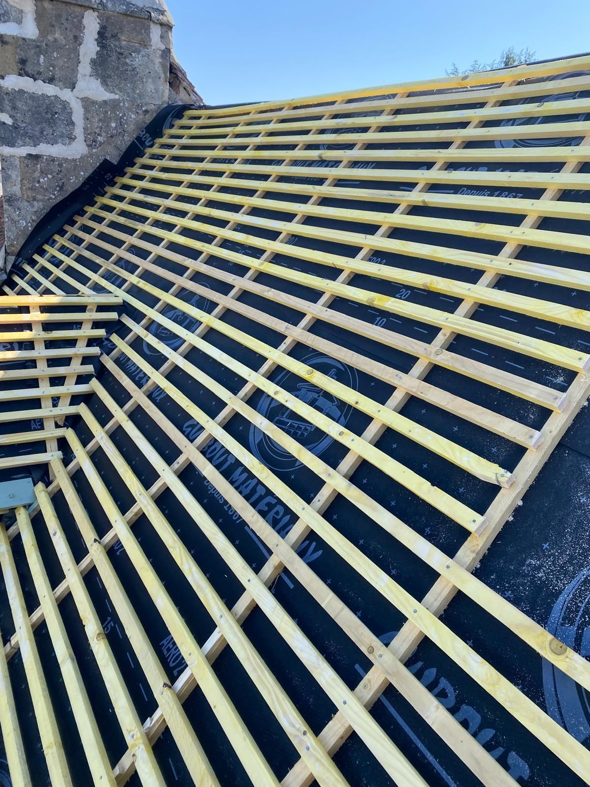 Charpente de toit en bois sur membrane d'étanchéité noire, près d'une structure en pierre.