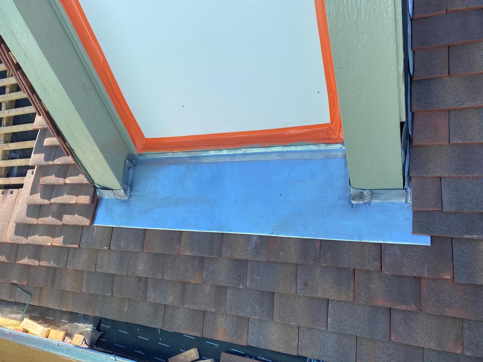 Gros plan sur un toit en bardeaux avec une fenêtre. On aperçoit les solins et les moulures, ainsi que la charpente en bois et le mastic orange.