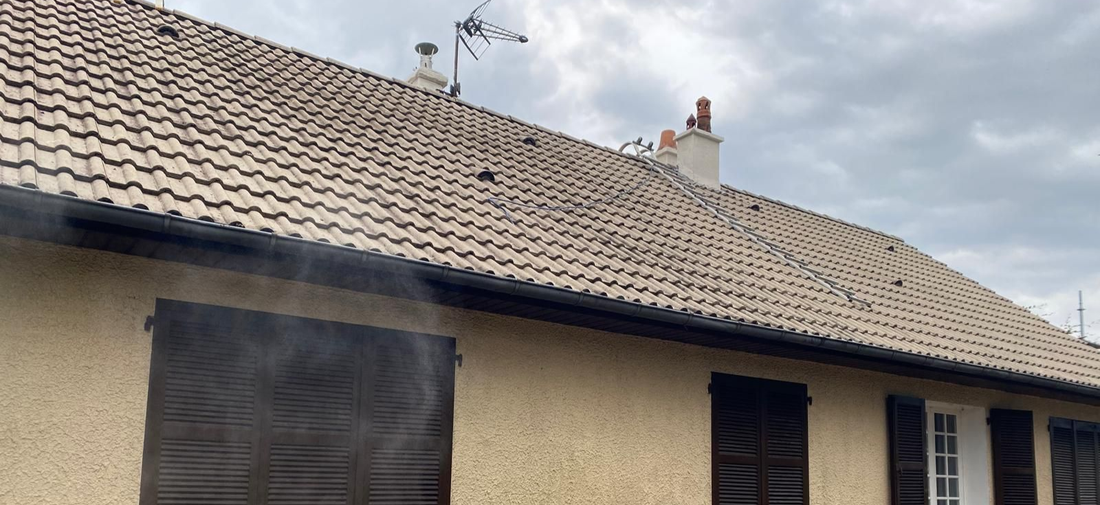 Une maison au toit de tuiles brunes et aux volets apparents, une cheminée et un ciel nuageux.