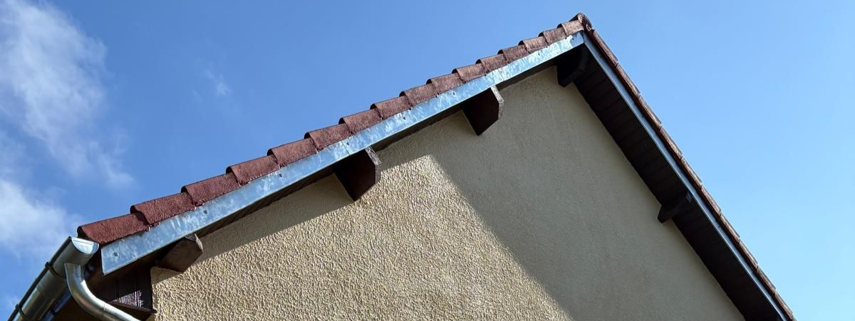 Vue de profil d'un toit de maison aux tuiles rouges et gouttière, se détachant sur un ciel bleu.