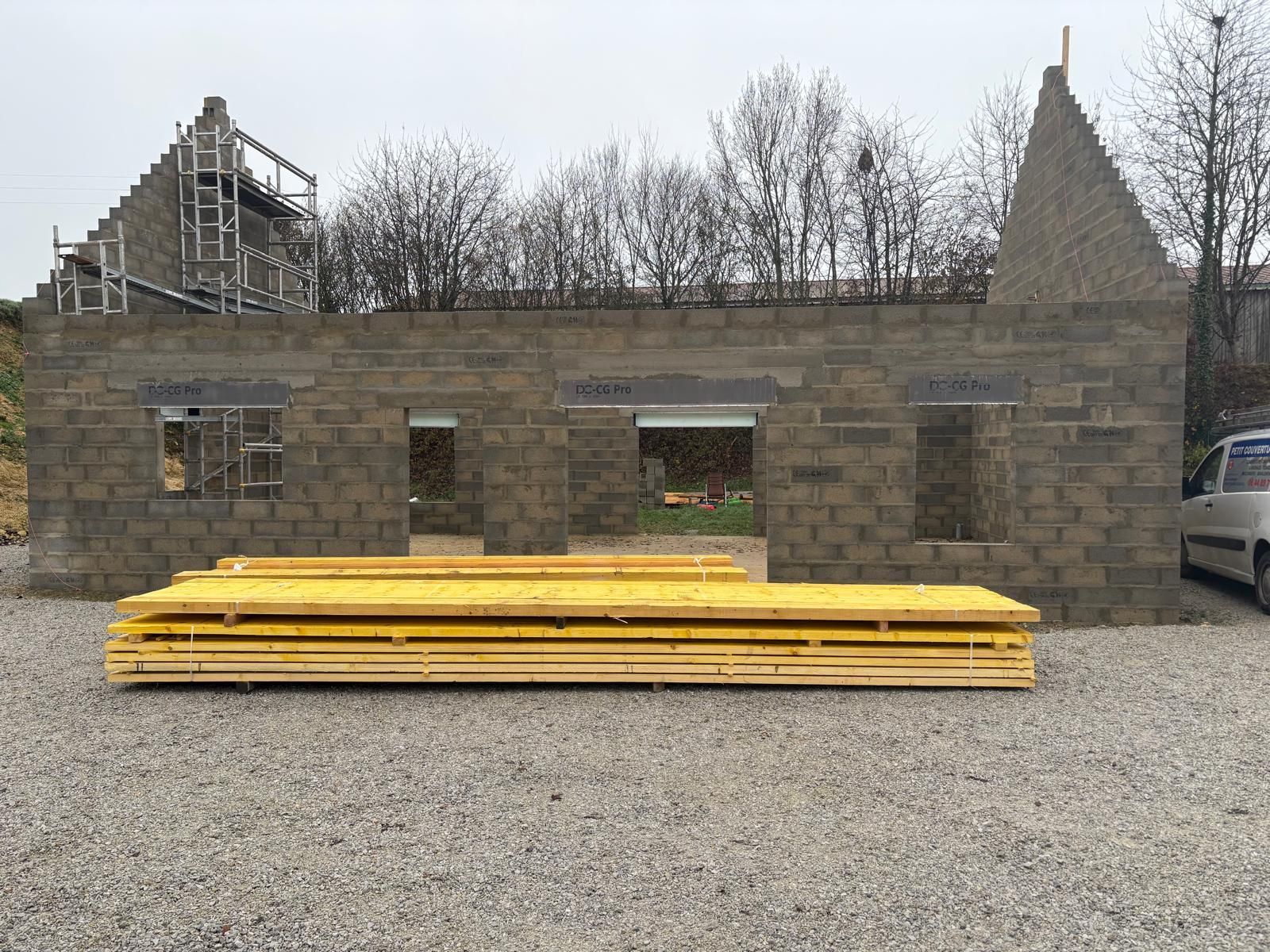 Bâtiment en construction ; murs en parpaings gris, bois jaune, sol en gravier et arbres en arrière-plan.