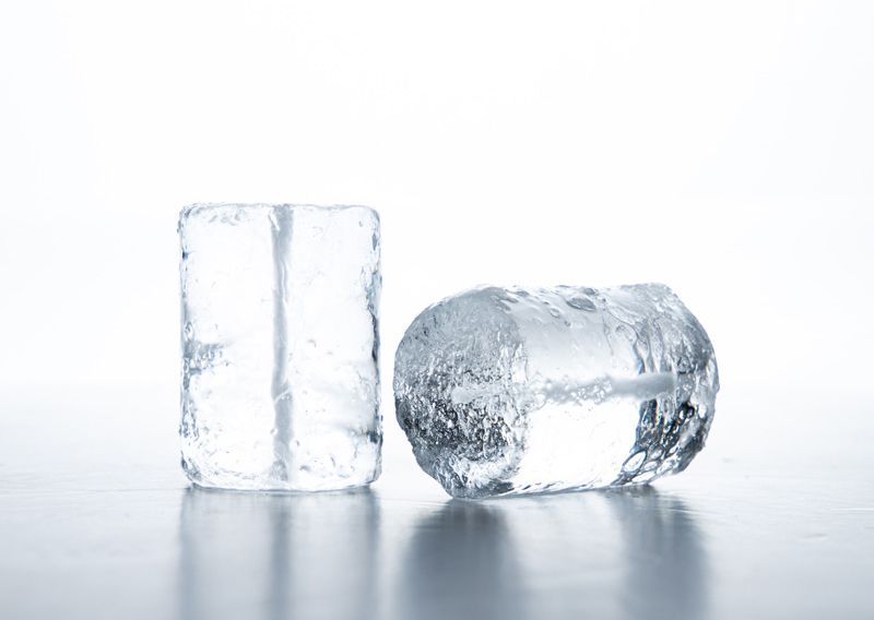 Dos cubitos de hielo, uno en posición vertical y el otro de lado, reflejando la luz sobre una superficie blanca.