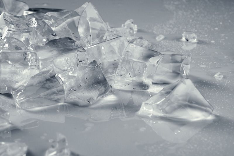Primer plano de cubitos de hielo rotos esparcidos sobre una superficie reflectante y de color claro.