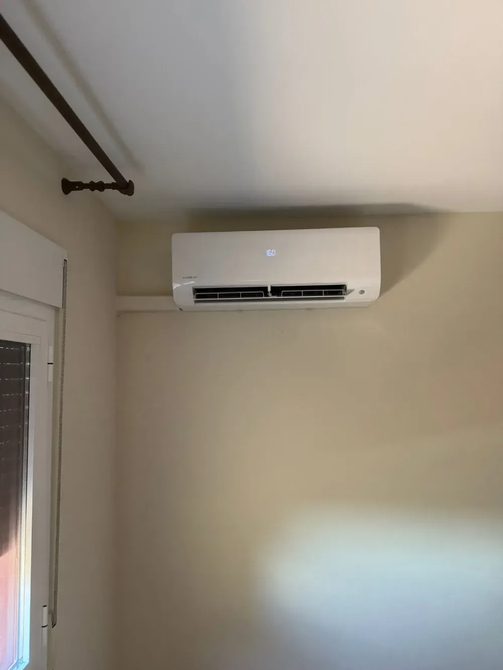 Aire acondicionado blanco montado en la pared, cerca del techo, en una habitación con una ventana y una barra para cortinas