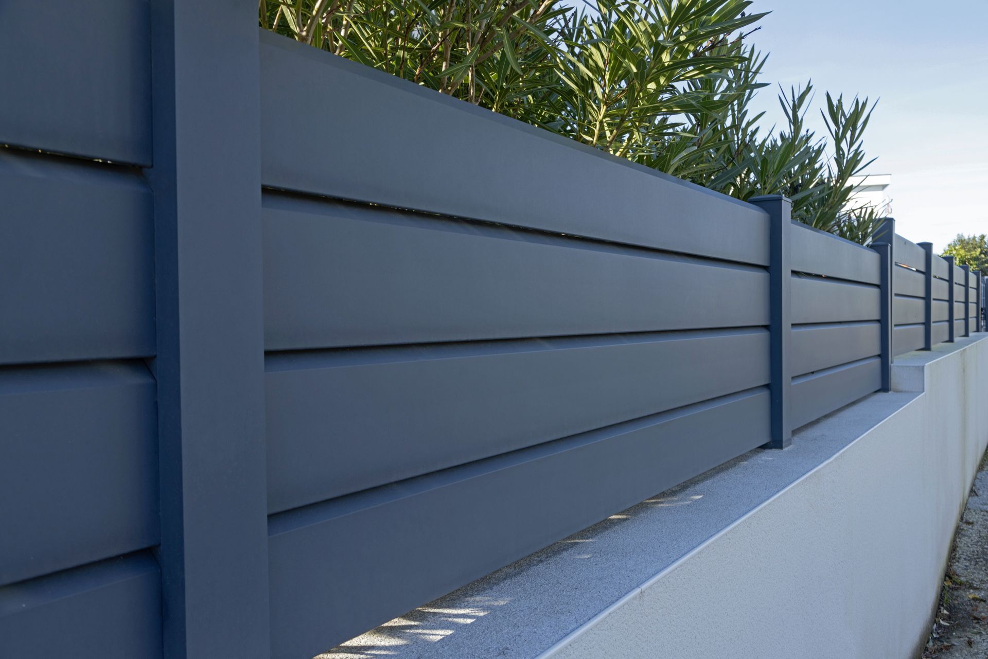 Une clôture moderne à lattes horizontales gris foncé, montée sur un muret en pierre claire, avec de la végétation verte en arrière-plan.