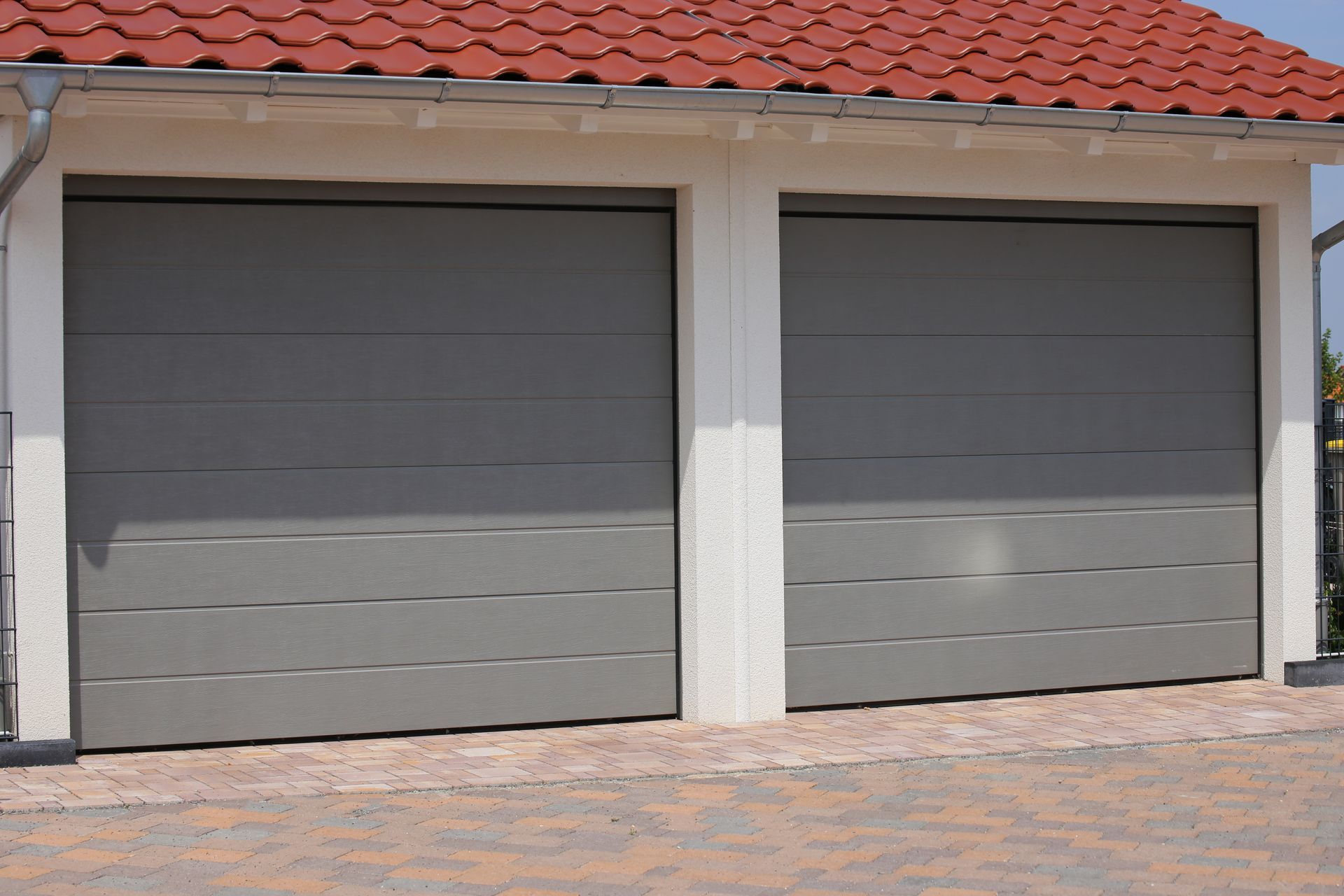 Deux portes de garage sectionnelles grises modernes installées côte à côte sur un bâtiment blanc au toit de tuiles en terre cuite.