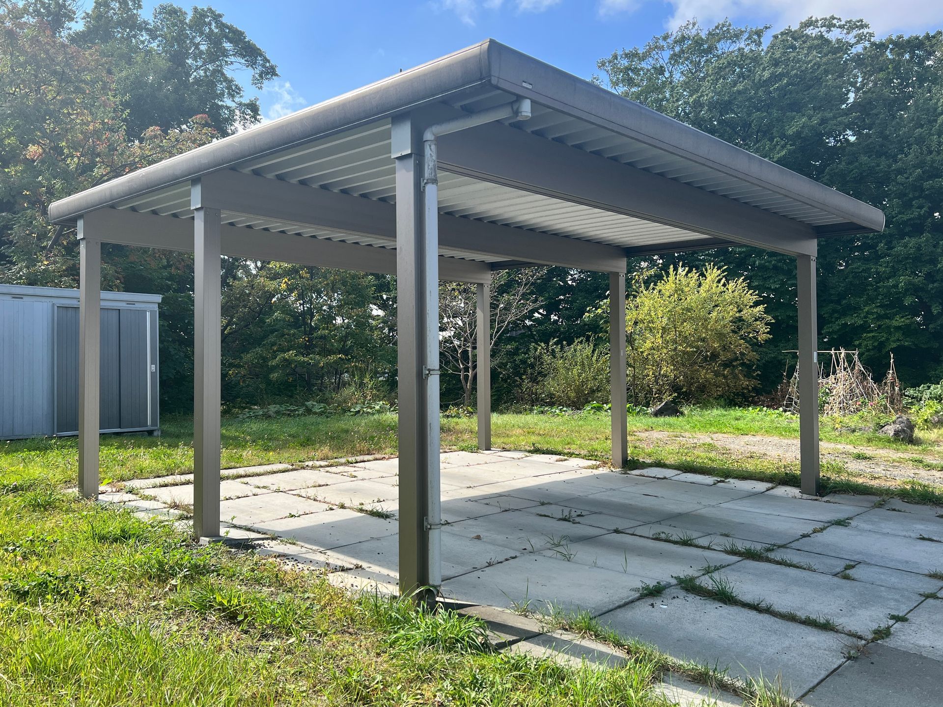 Un abri de voiture en métal brun se dresse au-dessus d'une dalle de béton, dans une zone herbeuse avec des arbres et un petit cabanon de rangement en arrière-plan.