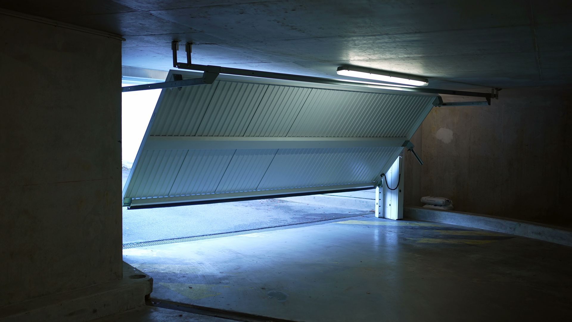 Une porte de garage blanche entrouverte, dévoilant un espace extérieur lumineux depuis l'intérieur d'un parking sombre en béton.