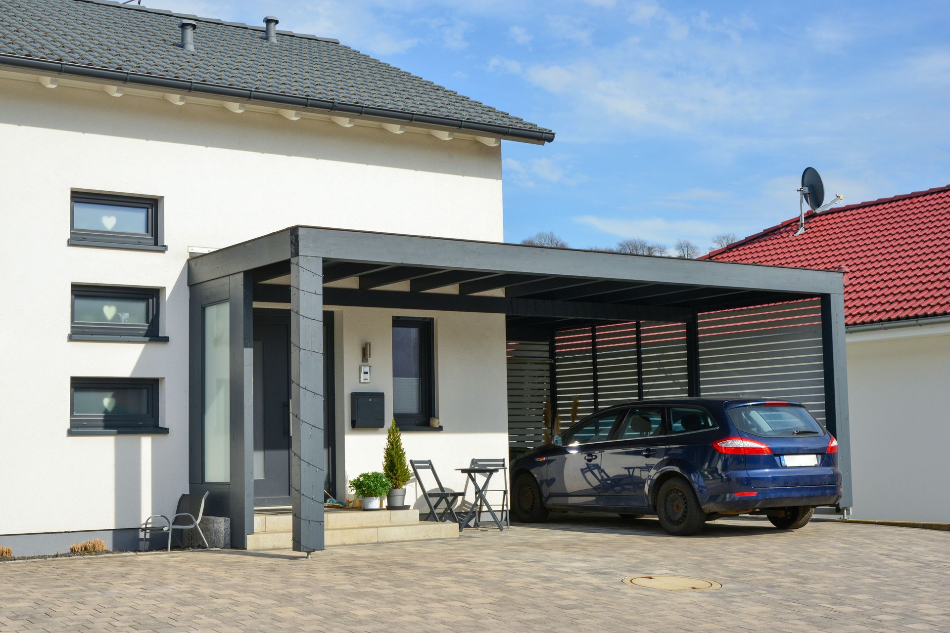 Une maison blanche avec un abri de voiture moderne gris foncé, avec un break bleu garé et un petit mobilier de terrasse.