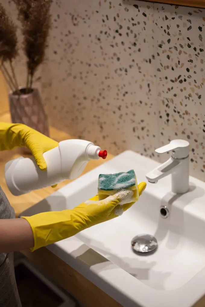 Persona con guantes amarillos limpiando un lavabo blanco con una esponja y una botella de limpiador en un baño.