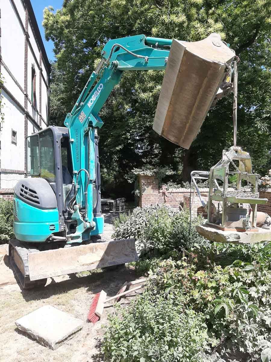 Ein blau-weißer Bagger trägt einen großen Holzblock - Traumgärten Benjamin Biermeyer aus Soest