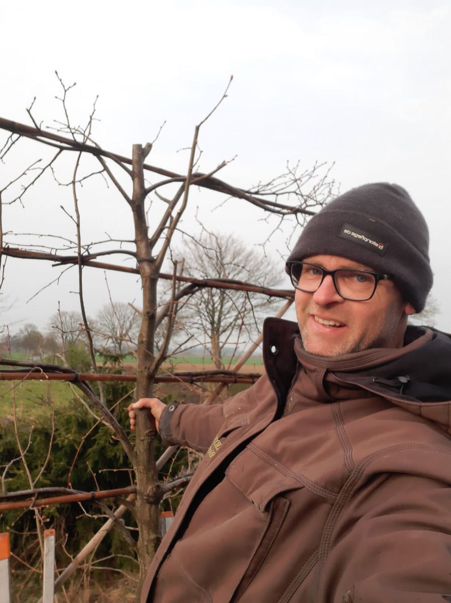 Ein Mann , der einen Hut und eine Jacke trägt, steht vor einem Baum - Traumgärten Benjamin Biermeyer aus Soest