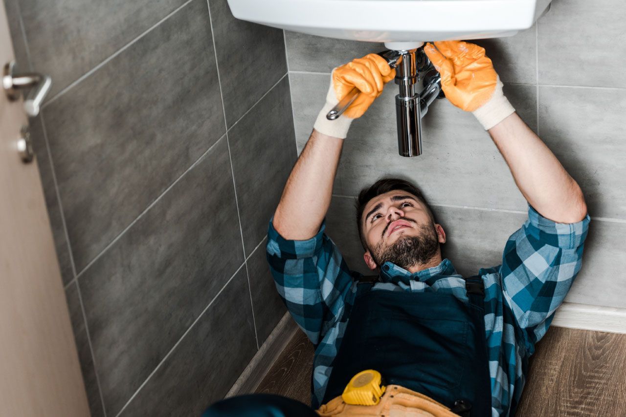Fontanero con guantes naranjas trabajando bajo un fregadero con una llave inglesa. Baño con pared de azulejos.