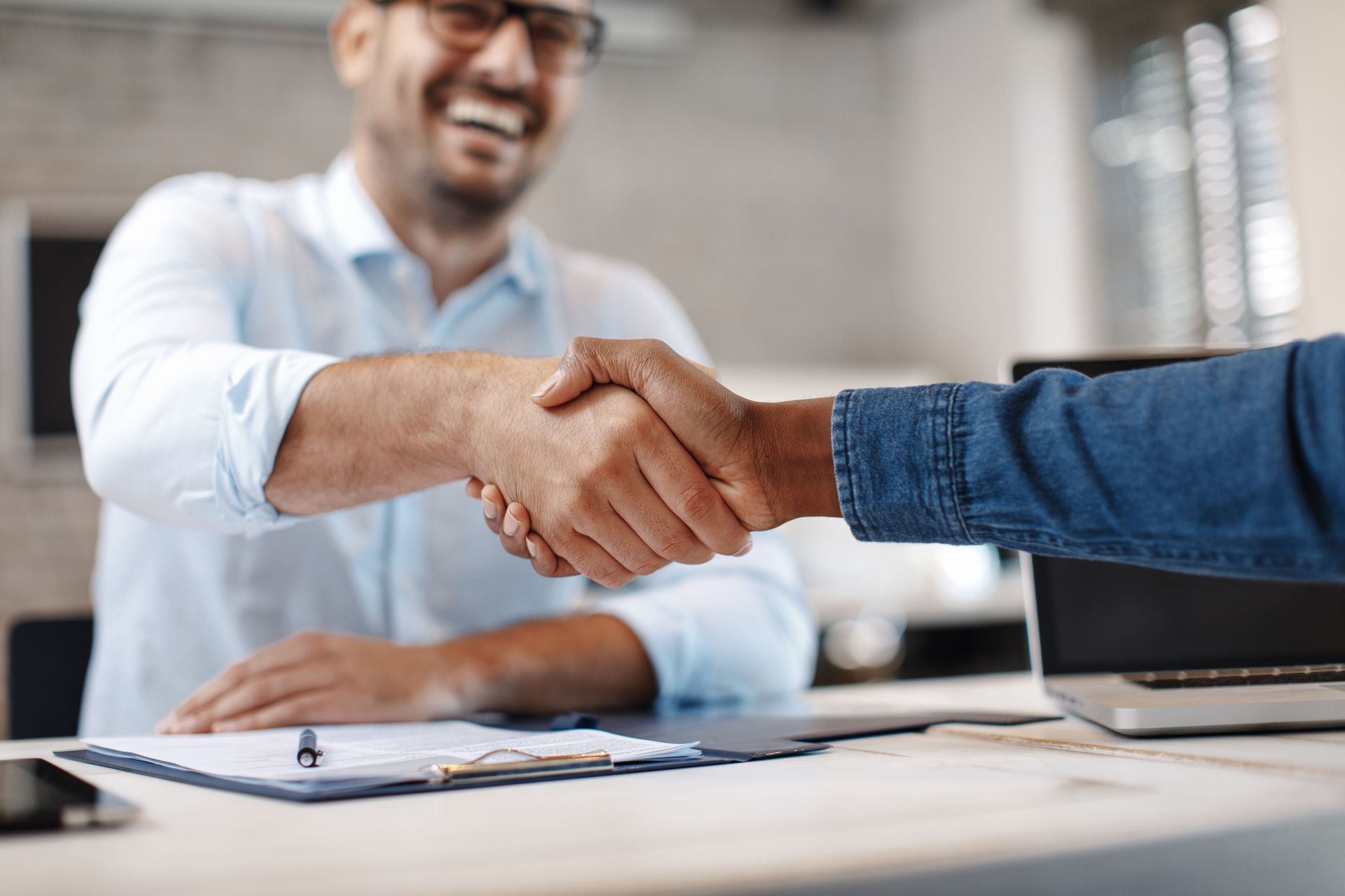Une poignée de main par-dessus un bureau avec une personne souriante, un bloc-notes et un ordinateur portable dans un bureau