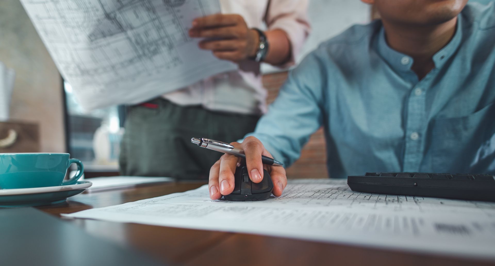Une personne écrit à un bureau, entourée de papiers et d'une calculatrice, tandis qu'une autre tient une feuille en arrière-plan.